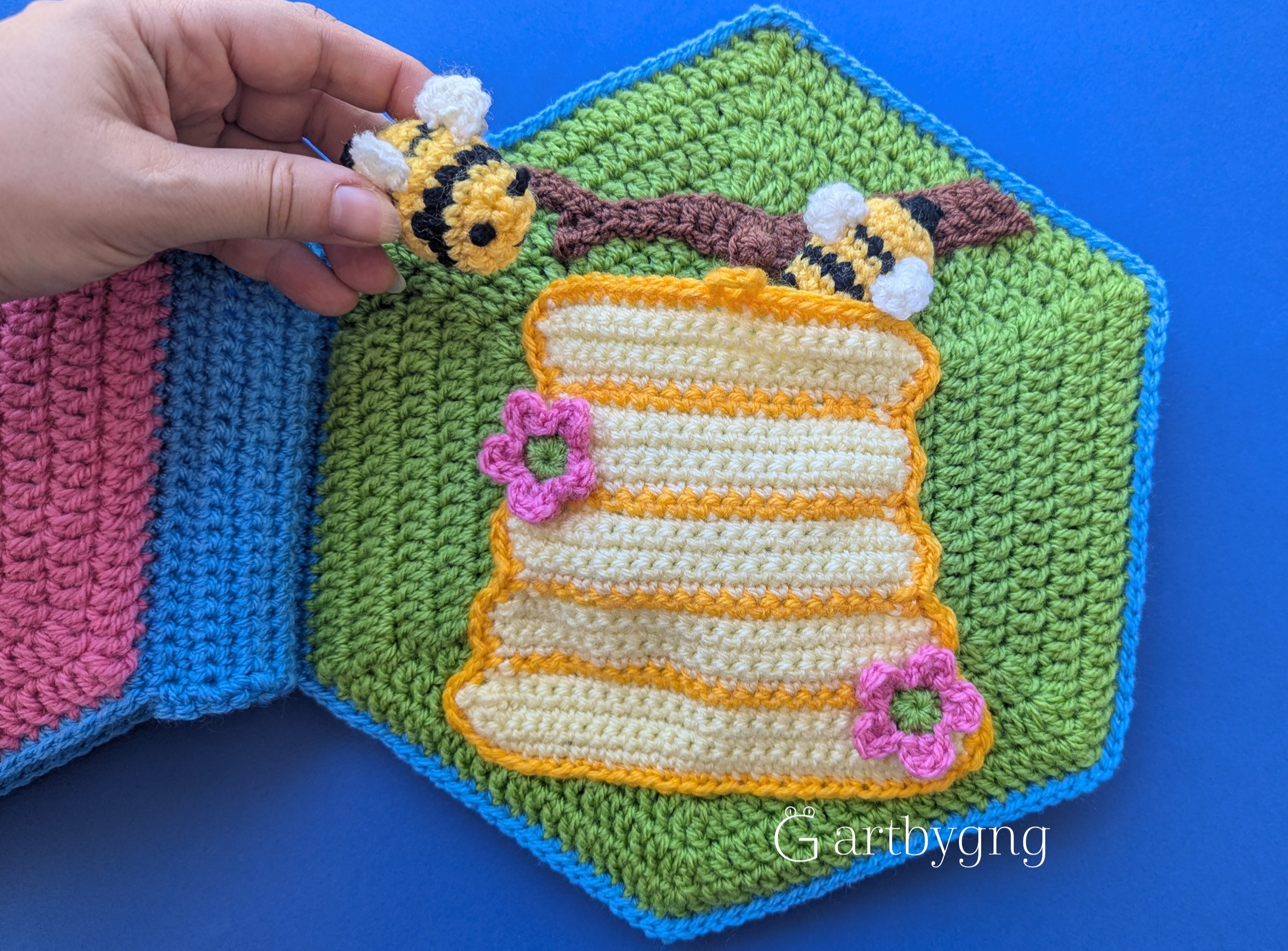 A hand holding a crocheted bee near flowers and a honeycomb hive pocket on a quiet book page