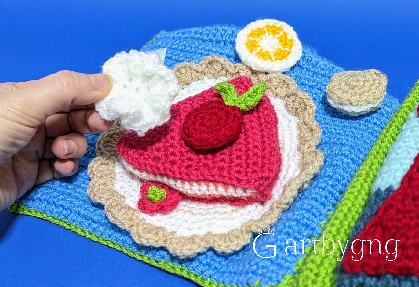 A hand placing a whipped cream topping onto a crocheted cake slice page, with a lemon slice and cookie piece nearby