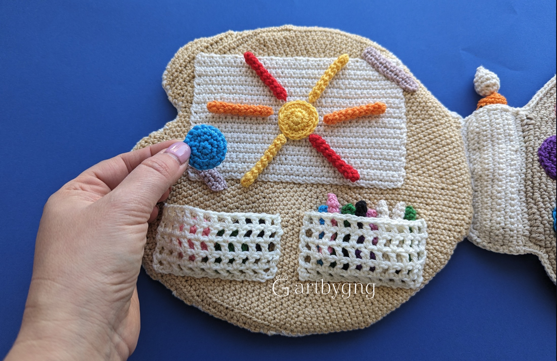 A hand placing a crocheted paint dot onto a palette-shaped page decorated with a sun design, with two mesh pockets holding extra paint pieces