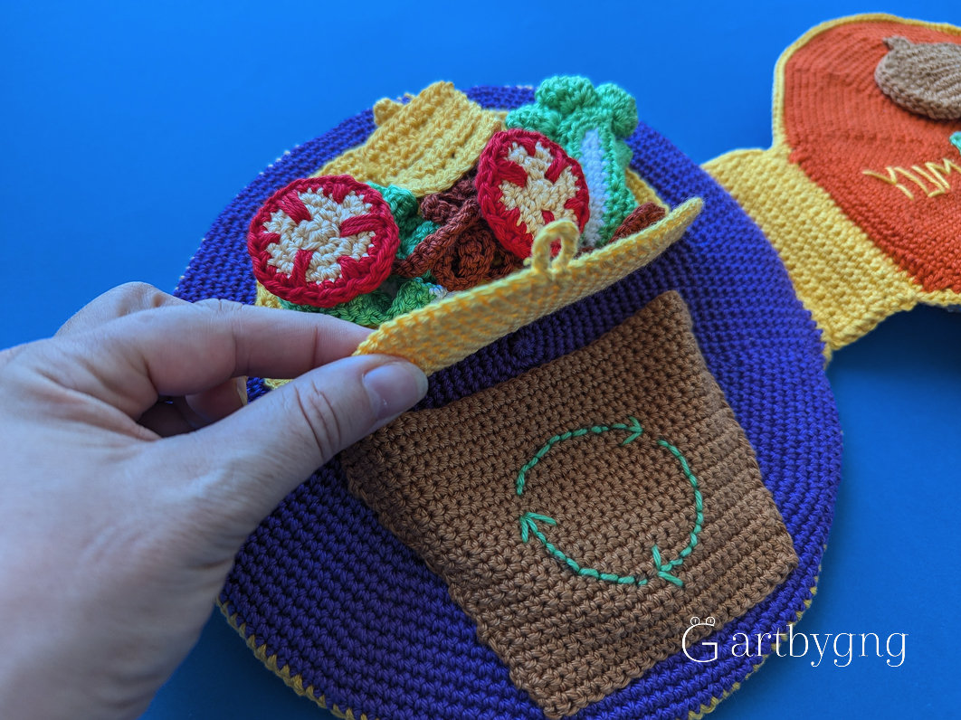 A hand lifting the flap of a crocheted taco-shaped pocket, with tomato, lettuce, and cheese play-food pieces tucked inside