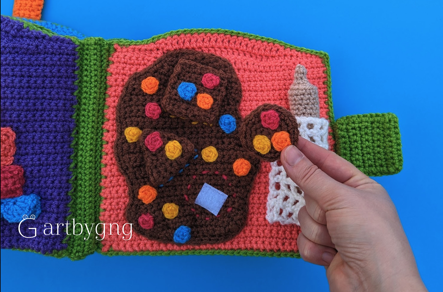 A hand placing a crocheted button-dotted shape onto a hand-shaped outline page, with a matching shape and milk bottle nearby
