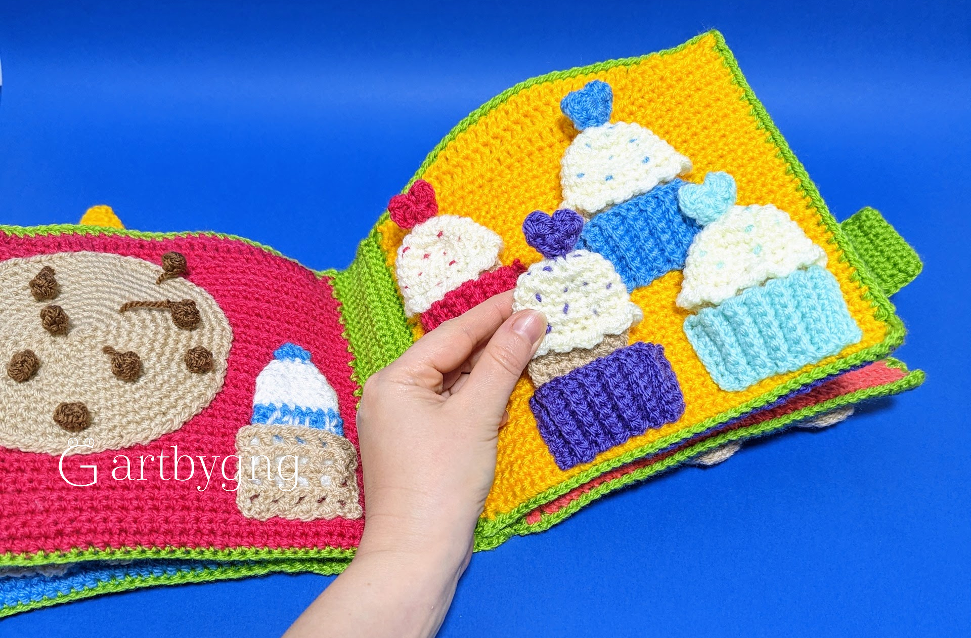 A hand picking up a crocheted cupcake piece from a busy book page, with cookie and toast pieces visible on the facing page